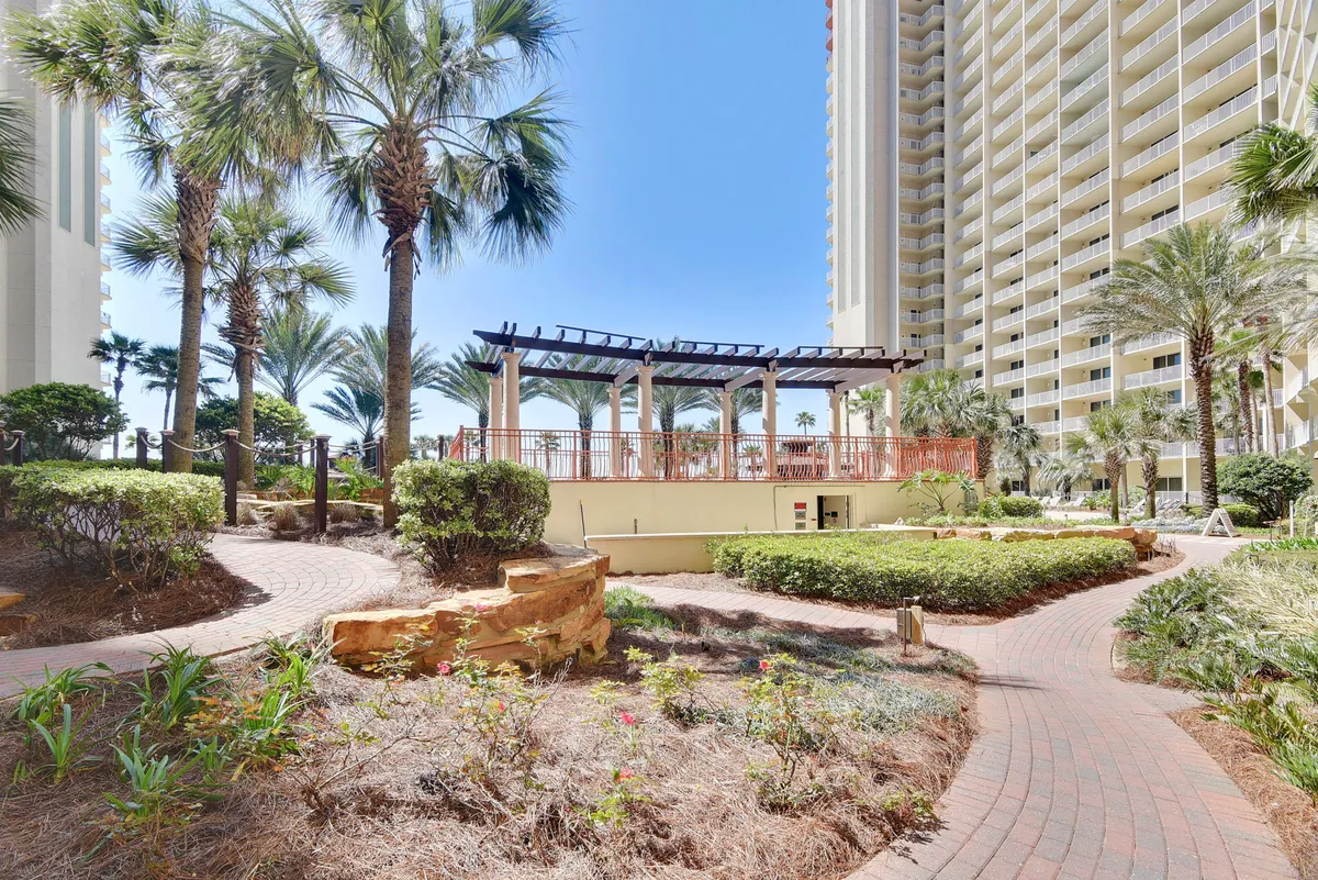 A serene landscaped garden with a pergola and winding pathway shaded by palm trees. The scene exudes tranquility, inviting guests to enjoy a morning coffee, stroll, or gather outdoors with friends in a peaceful setting.