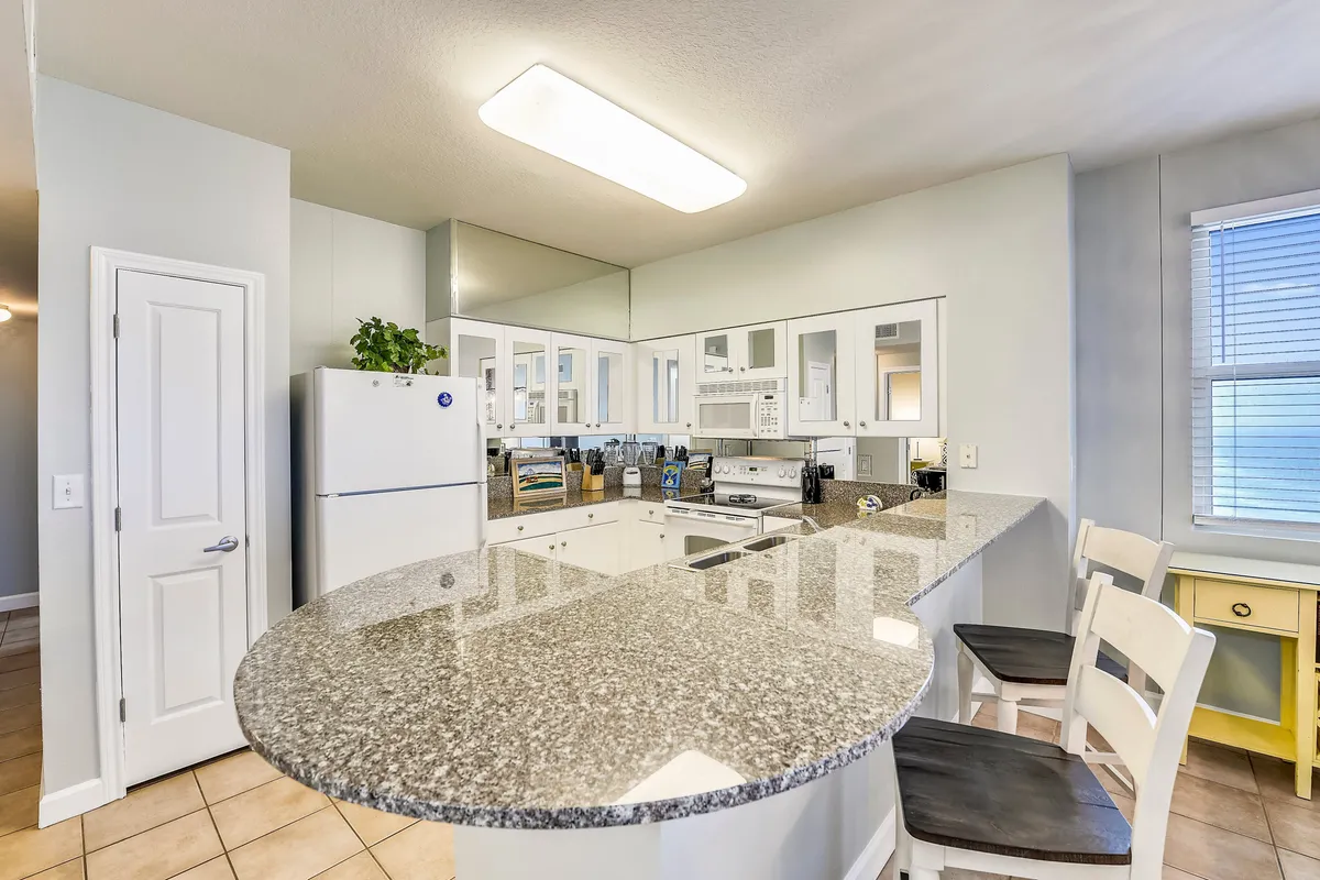 A bright, modern kitchen highlighted by a spacious granite countertop, sleek white cabinetry, bar stools for casual seating, and high-end kitchen appliances. The scene is bathed in light, suggesting a lively and welcoming environment ideal for meal prep and informal gatherings.
