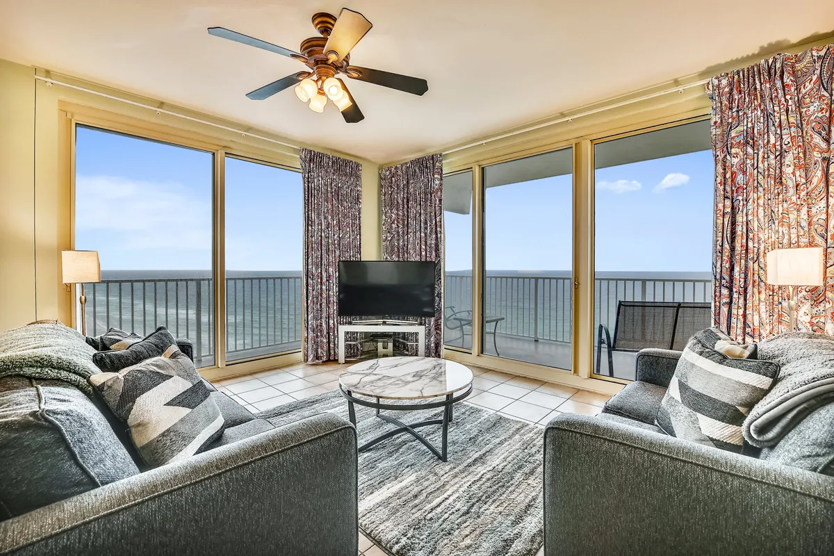 A cozy, sunlit living room with large windows framing a stunning ocean view. The space features comfortable seating, decorative curtains, a coffee table, and soft lighting, creating an inviting atmosphere perfect for relaxing, watching TV, or hosting gatherings.