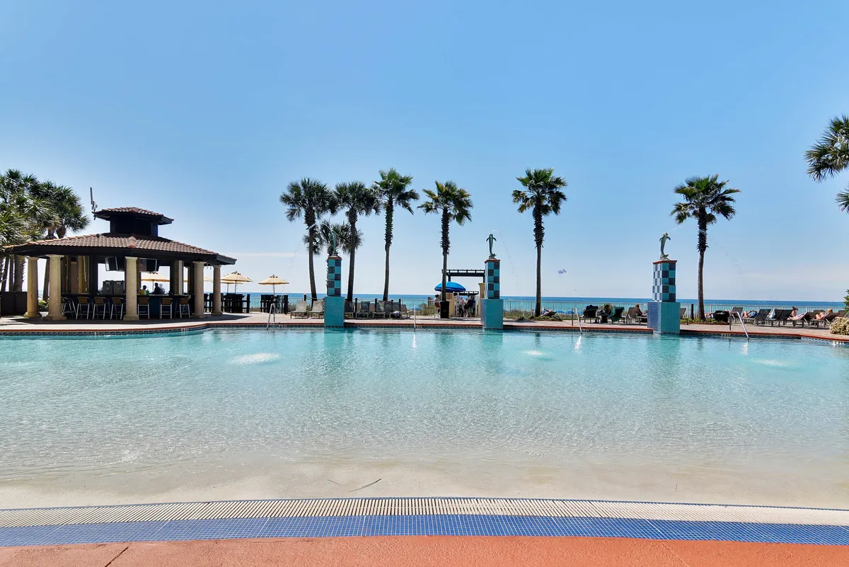 This image captures the inviting lagoon-style swimming pool surrounded by palm trees, tiki bar, lounge chairs, and, just beyond, a sandy beachfront and gentle Gulf waves. Families are seen enjoying the pool and the adjacent beach, creating an atmosphere of fun, relaxation, and tropical excitement.