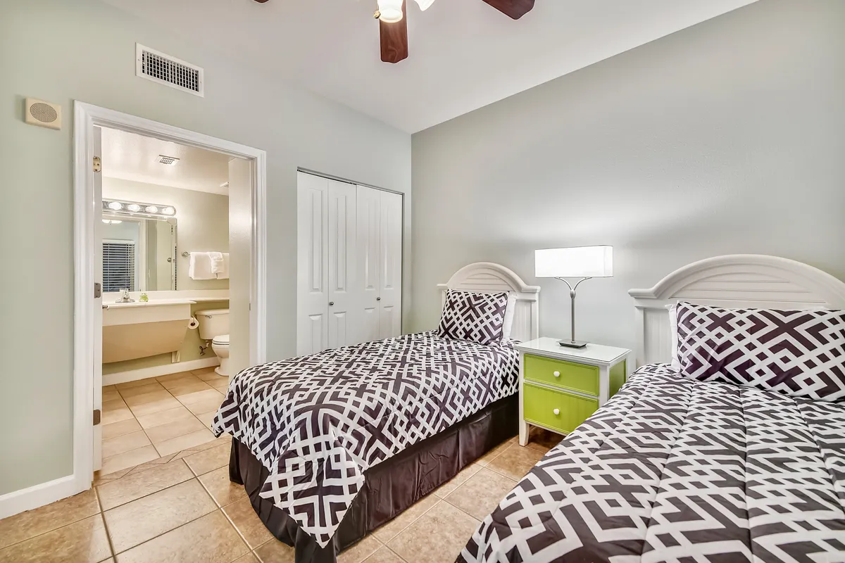 A twin bedroom with two beds adorned in patterned bedding, a green nightstand with a lamp between them, and an open doorway leading to a nearby bathroom. The room exudes a relaxed, welcoming mood, ideal for children or friends.