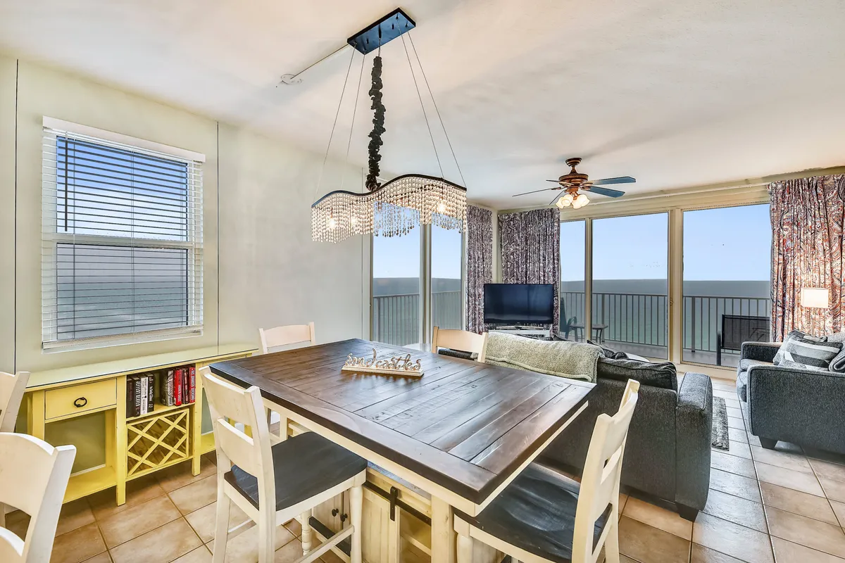An open-concept living area with a large wooden dining table, white chairs, and an elegant crystal chandelier. The space features floor-to-ceiling windows that frame stunning Gulf views. The comfortable living area with a sofa and TV is adjacent to a full-sized kitchen, all bathed in natural sunlight.