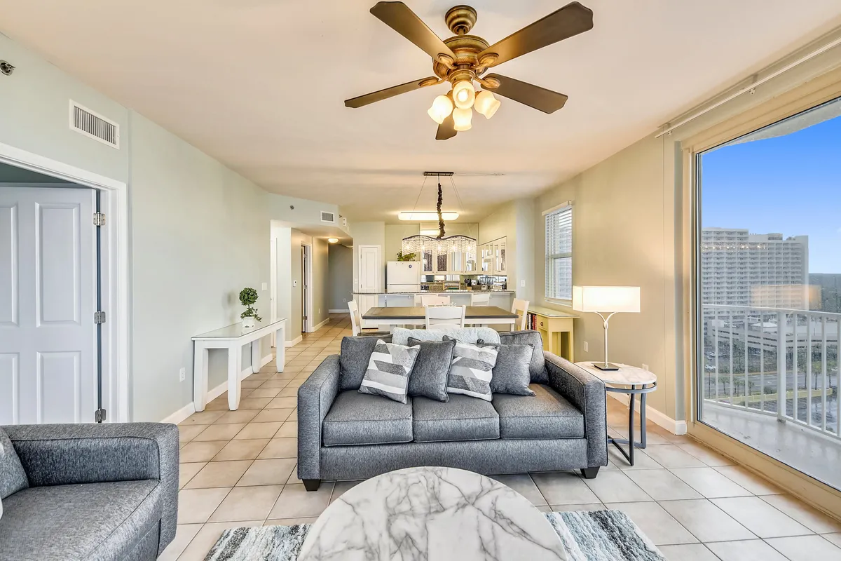 A modern, comfortable living space with a gray sofa, patterned pillows, and a marble coffee table faces floor-to-ceiling windows that offer a balcony view. The open layout connects to the kitchen and dining area, filled with natural light and a fresh coastal feel.