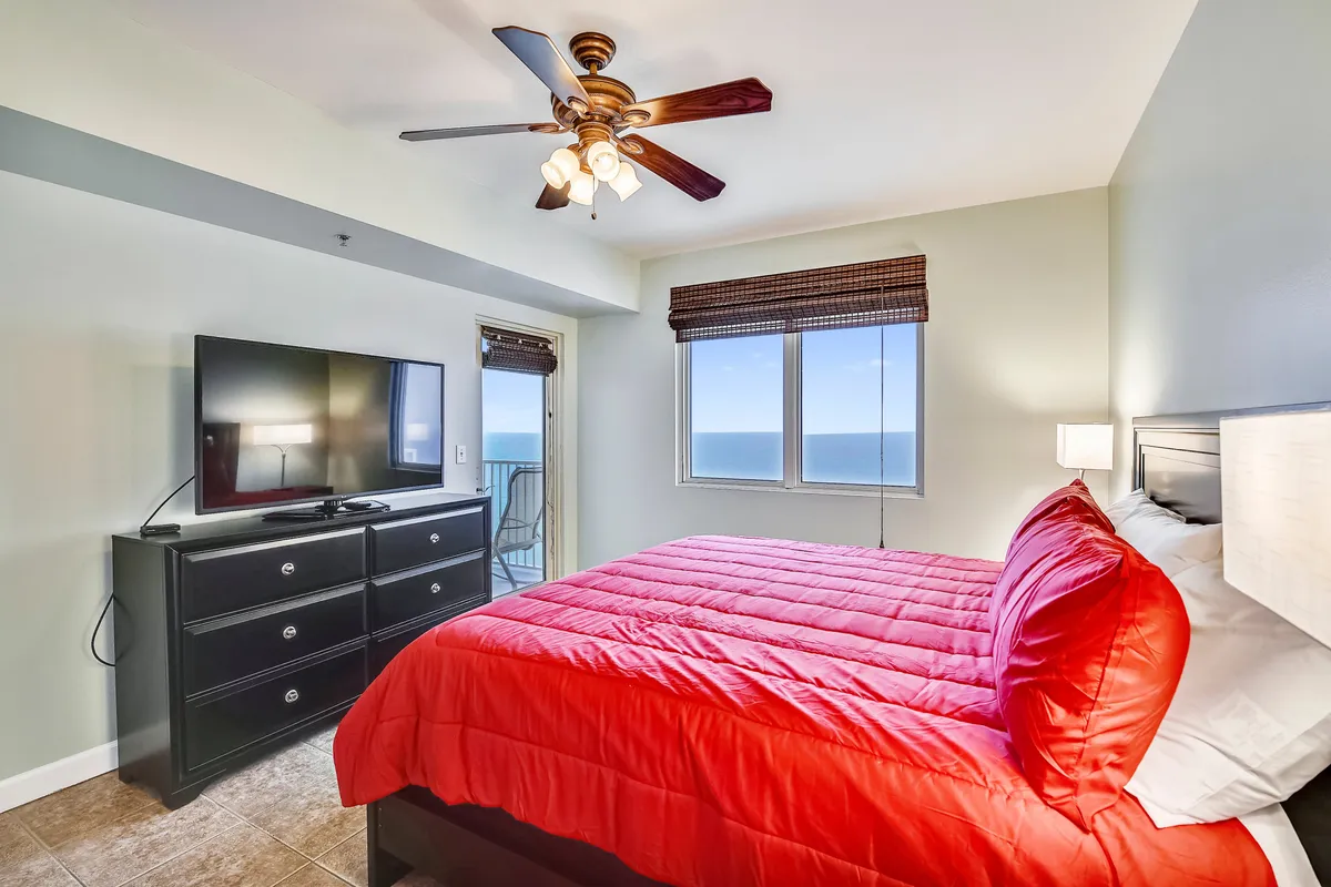This image presents a master bedroom with a king size bed adorned in bright red bedding, a large window offering Gulf views, a sleek black dresser with a flat screen TV, and a ceiling fan with lights. The room is spacious and filled with natural light, creating an inviting and relaxing ambiance.