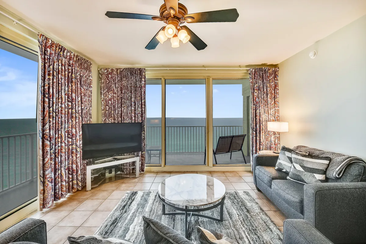 A bright, airy living room with floor-to-ceiling windows offering expansive ocean views. The space features a comfortable grey sofa, striped cushions, a round marble coffee table, and a flat screen television, with the balcony just beyond the windows.