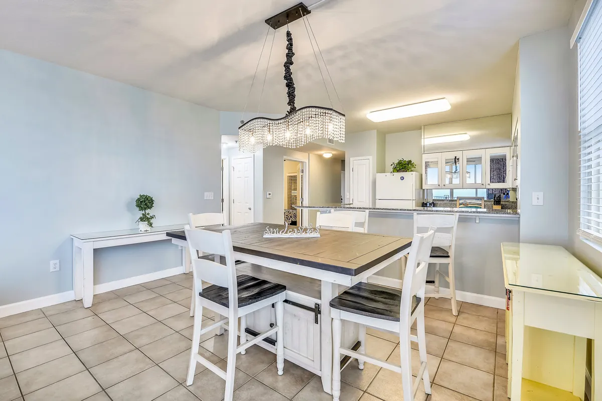 A bright, inviting open-plan kitchen and dining area features a wooden table with chairs, a unique chandelier, a breakfast bar, and a 'sandcastles' sign as a playful beach-inspired detail. The kitchen is fully equipped for meal preparation and family gatherings.