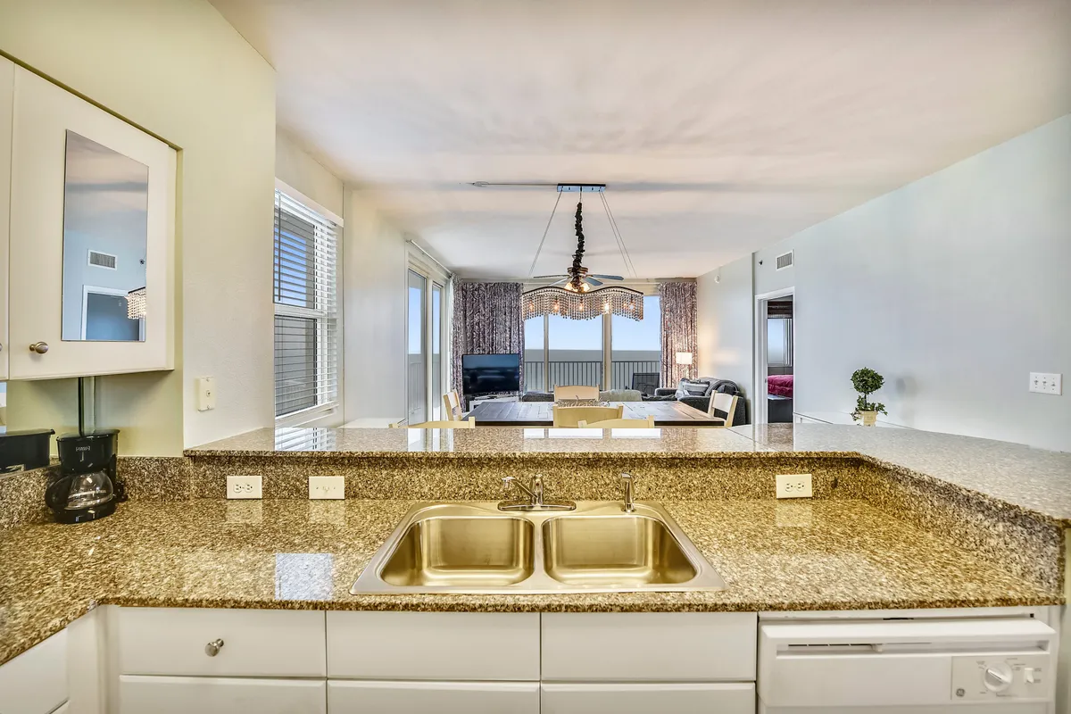 A modern open-concept kitchen overlooking a living and dining area, with floor-to-ceiling windows that flood the space with sunlight and offer direct views of the Gulf. The granite countertop, modern chandelier, and dining set are all visible, creating a sense of cohesive, upscale beach living.