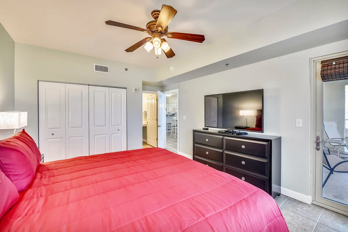 A bedroom with a bright red bedspread and pillows, a ceiling fan with lights, a flat screen TV on a black dresser, and a door leading to a balcony with outdoor chairs visible. The mood is cozy and restful, with easy access to fresh air and views.