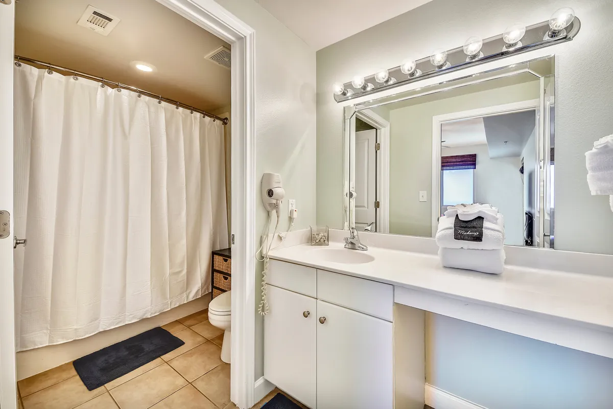 A fresh, organized bathroom with a large mirror, bright vanity lights, white countertop with folded towels, a shower area with a white curtain, and a wall-mounted hairdryer. The space is clean and inviting, perfect for self-care routines.