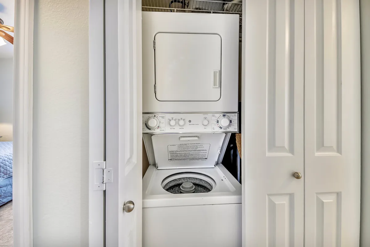 A practical laundry area with a stacked washer and dryer unit is tucked behind white bifold closet doors. The scene also offers a glimpse of a bedroom with blue bedding, reinforcing the home's residential comfort.