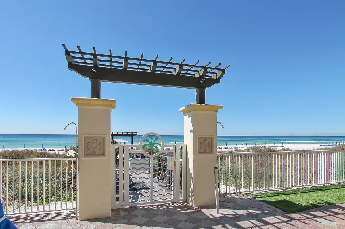 This image captures a decorative entrance gate adorned with palm and wave motifs, opening to reveal pristine white sand, turquoise Gulf waters, and a row of blue beach umbrellas along the shore. The clear blue sky stretches across the horizon, creating a sense of openness and escape.