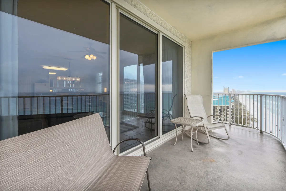 A luxurious wraparound balcony with seating, curved white railing, and floor-to-ceiling sliding glass doors. The balcony offers panoramic views of the Gulf and beachfront, providing outdoor space for relaxation or social gatherings.