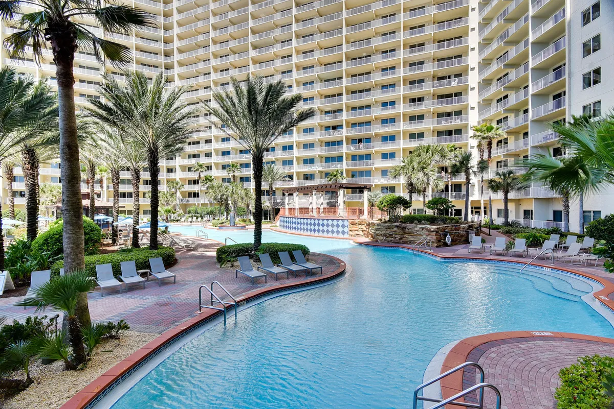 A large lagoon-style swimming pool with fountains is surrounded by tropical palm trees, a tall resort building with multiple balconies, and rows of poolside lounge chairs. The inviting pool deck is spacious, offering plenty of room for families to relax, swim, and socialize in a lush, resort-like environment.