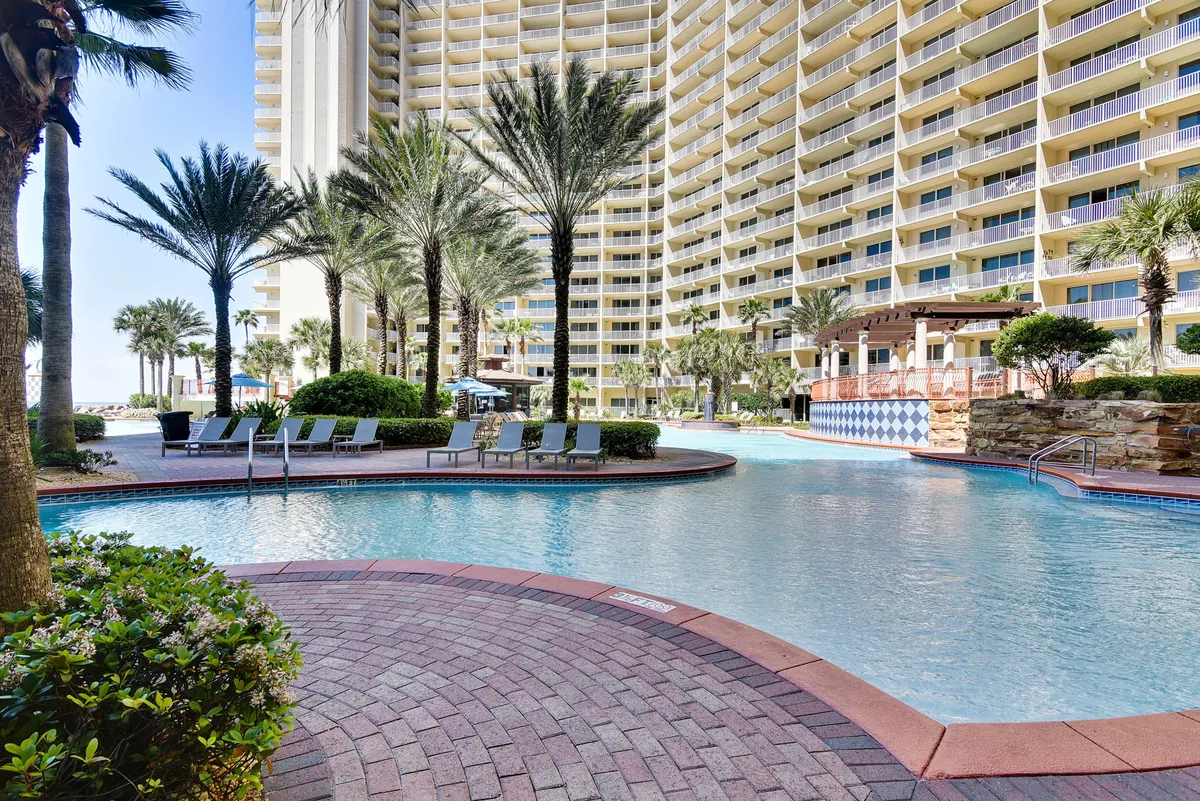 A sprawling lagoon-style swimming pool sits at the heart of the resort, surrounded by tall palm trees and a multi-story resort building with balconies. Poolside seating areas and a shaded pergola offer spots to gather and relax, while guests can be seen swimming and socializing. The lively, sunlit scene evokes vacation fun and resort energy.