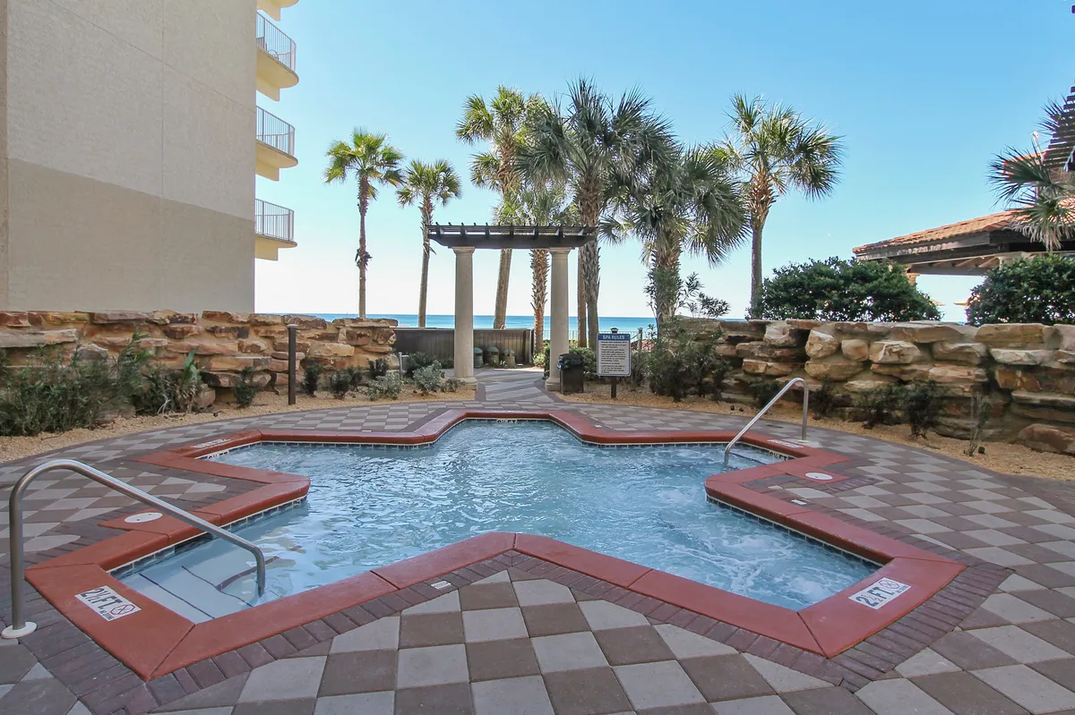 A star-shaped hot tub bubbles invitingly amidst tropical landscaping, with palm trees framing an open view toward the Gulf. The stone wall and lush greenery create a private, spa-like ambiance, while the ocean breeze is suggested by the open vista.