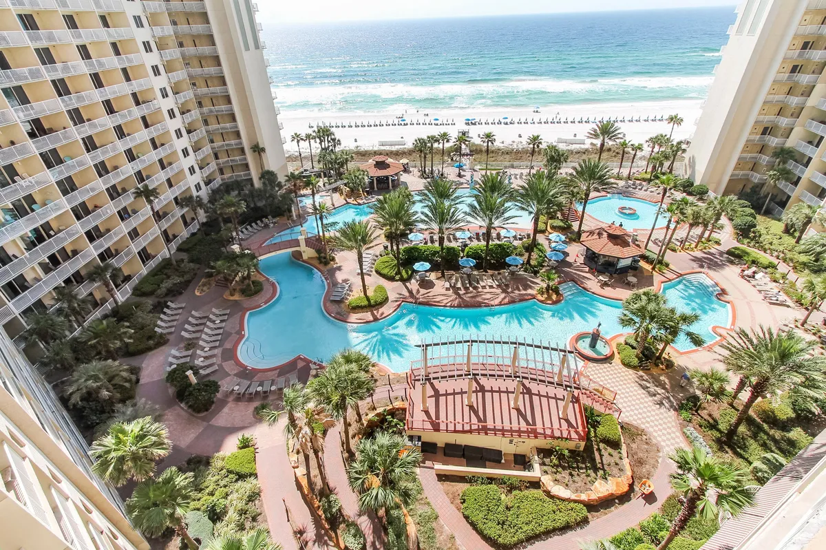 A sprawling lagoon-style swimming pool is surrounded by lush palm trees and tropical landscaping. The pool area features shaded tiki bars and opens directly onto a white sandy beach with the turquoise Gulf of Mexico visible in the background. Lounge chairs line the pool deck, inviting relaxation and family fun.