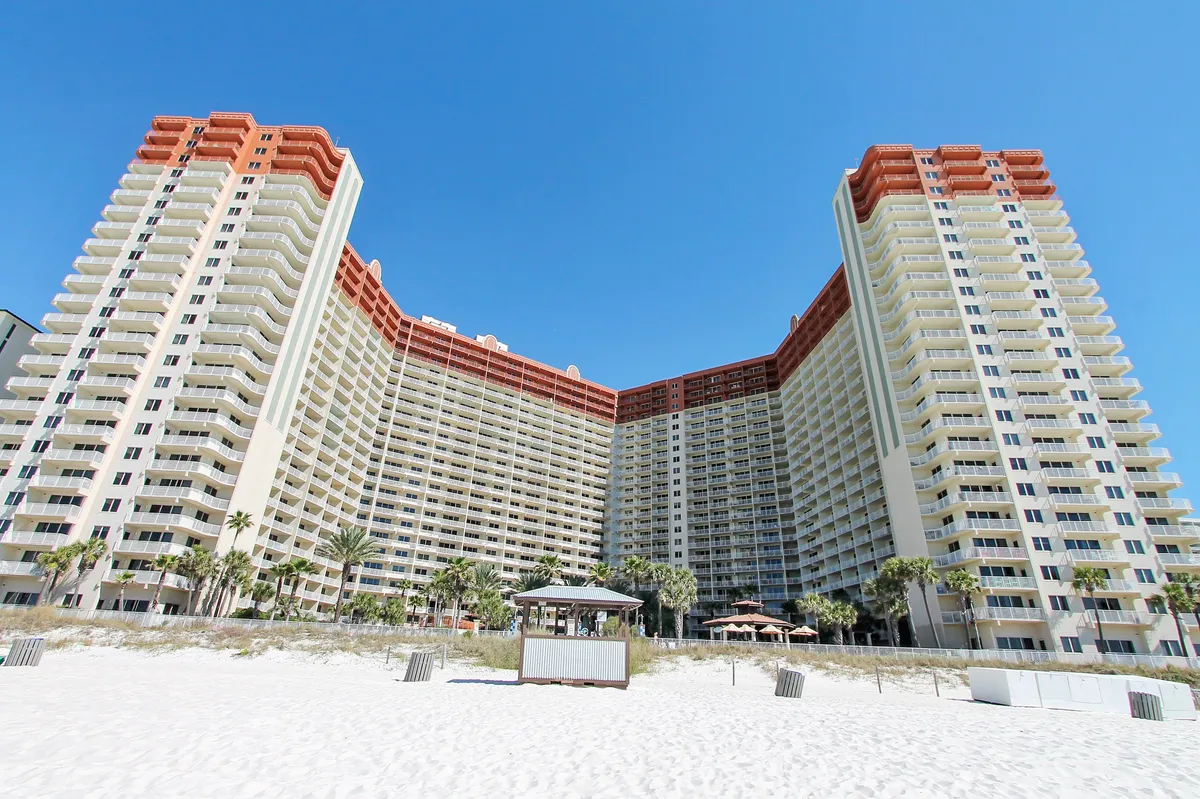 A vibrant exterior shot of a large lagoon-style swimming pool set against a multi-story resort building, with poolside cabanas, umbrellas, and the white sandy beach visible in the foreground. The inviting pool is the centerpiece, surrounded by palm trees and tropical landscaping, reflecting an upscale, leisurely atmosphere.