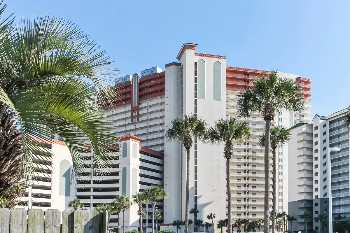 An exterior shot featuring a tall resort condominium with distinctive red roof accents, framed by clusters of palm trees and vibrant blue sky. The lush landscaping and tropical plants create an inviting, sun-soaked resort atmosphere.