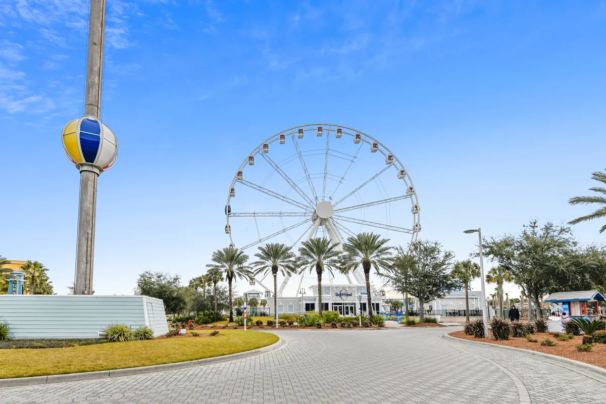A large Ferris wheel rises above palm-tree-lined walkways, with a colorful balloon-shaped structure and a curved paved path creating a lively, cheerful scene. The setting suggests proximity to major attractions and opportunities for shared adventures beyond the resort.