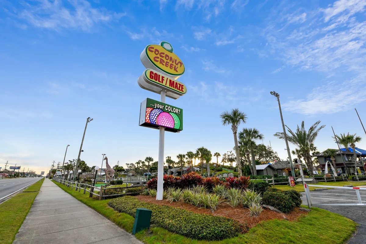 A colorful sign marks the entrance to Coconut Creek Golf/Maze, with a miniature golf course visible behind palm trees and lush landscaping. The cheerful, open-air space is designed for playful competition and outdoor family bonding.