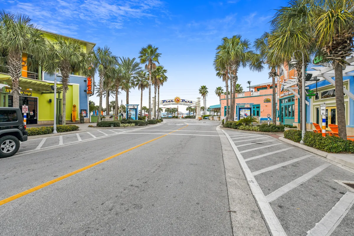 Palm trees line a lively street of colorful storefronts beneath a blue sky, with an archway reading 'Pier Park.' The vibrant setting suggests nearby dining, shopping, and entertainment options, evoking the energy of the local area just steps from the resort.