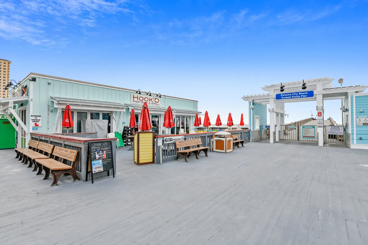 The Hook'd Pier Bar and Grill sits at the entrance to the Panama City Beach Russell Fields Pier, with inviting red umbrellas, benches lining the boardwalk, and the ocean visible in the background. The image captures people enjoying the casual, social vibe and easy access to dining and nightlife, all steps from the beach.