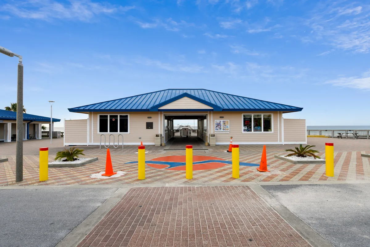 A welcoming exterior perspective of the beach access building, featuring a blue metal roof and a decorated pathway leading through the entrance directly to the beach. The pavement sports a colorful fish emblem, and the scene captures the anticipation and ease of reaching the shoreline.