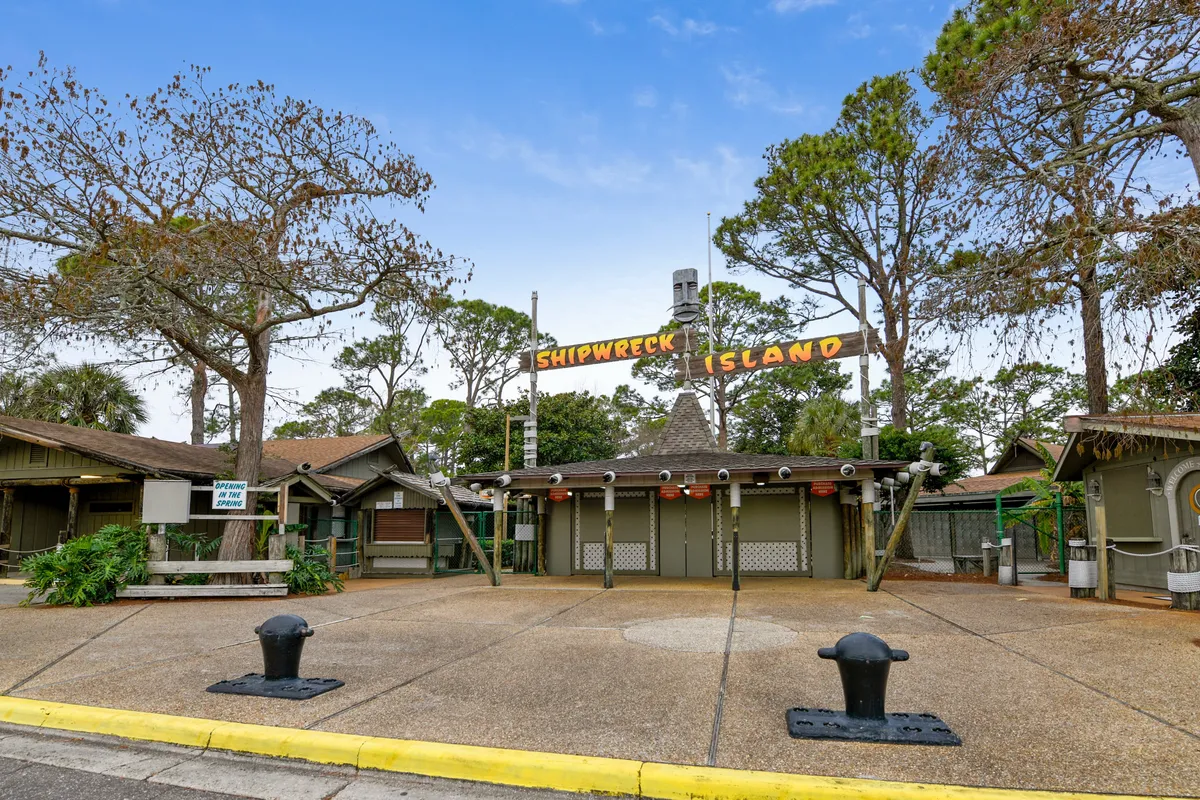 The entrance to Shipwreck Island Water Park features a rustic wooden sign and structures, with a large paved open area and trees framing the scene. The image exudes anticipation for thrilling water park adventures and highlights one of the complimentary local attractions available to guests.